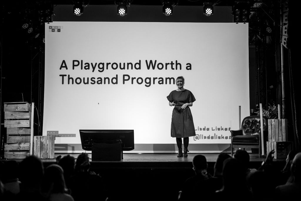 A black and white photo of Linda Liukas smiling massively from the stage into the audience at beyond tellerrand in Berlin 2024. She wears a dress. The slide on the canvas in her back shows the title of her talk “A Playground Worth a Thousand Programmes”