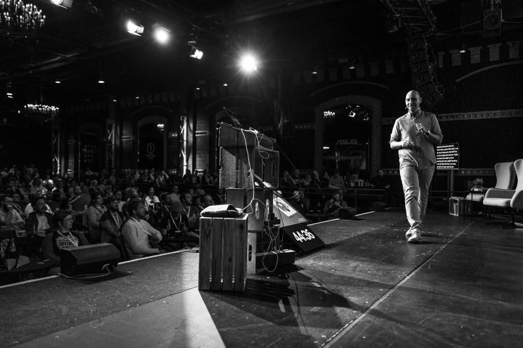 A black and white photo of Vitaly Friedman on stage at beyond tellerrand. The photo is taken from the side of the stage and you can see a packed room on the left side of the photo with attendees following the presentation.