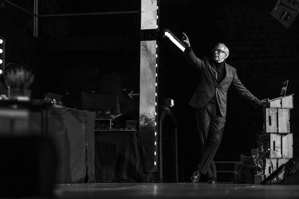 A photo of Brendan Dawes during his talk which was taken from the left side of the stage. Brendan has turned his back to the audience as he points to something on the canvas.