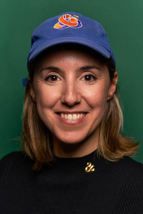 Portrait of Martina Flor wearing a blue baseball cap with a red ampersand logo. Shot in front of a green background.