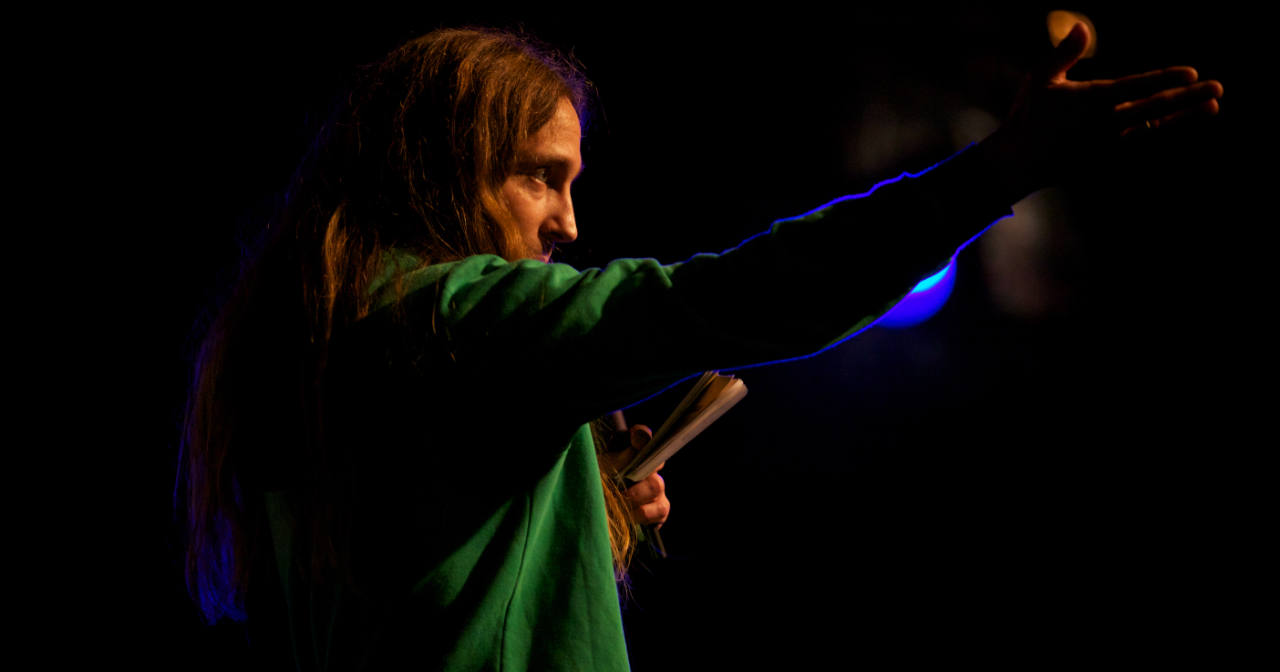 The founder of beyond tellerrand, Marc Thiele, in a close-up from the side in front of a blue light. He has long hair and wears a green hoodie and his arm makes a gesture as if he s explaining or showing something to the audience. His other hand holds a microphone and a pocket notebook.