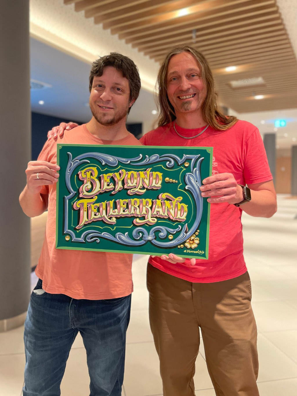 A photo that show Gustavo handing over the finished painting to Marc. Both holding the sign into the camera together smiling happily.