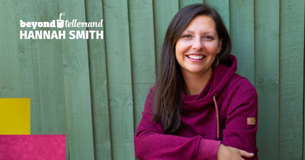 A photo of Hannah Smith in front of a green wooden wall or fence. She is smiling into the camera and wears a pink hoodie.