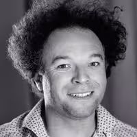 A portrait photo of Jude Pullen. He is a man, has curly wild hair and smiles into the camera.